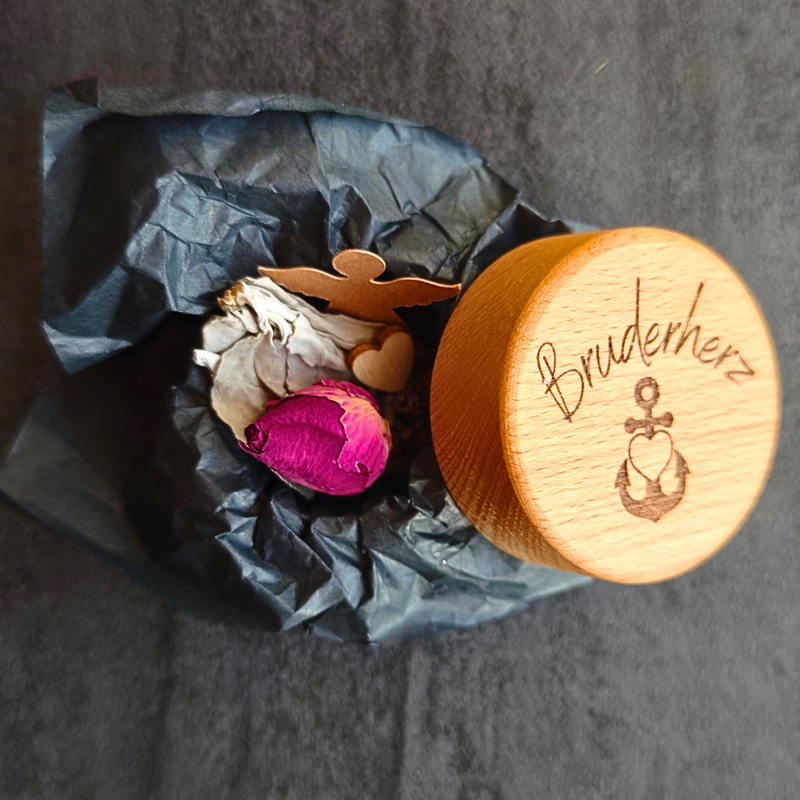 Geöffnete Mementovia-Box "Stille Umarmung" mit den symbolischen Beigaben Lavendel, Rose, Salbei, Engel und Herz – sanfte Begleiter in der Trauer. Im Vordergrund der Deckel der Box mit dem Motiv "Glaube, Liebe, Hoffnung" und dem gravierten Wort "Bruderherz" - Die Beigaben liegen auf schwarzem Seidenpapier.