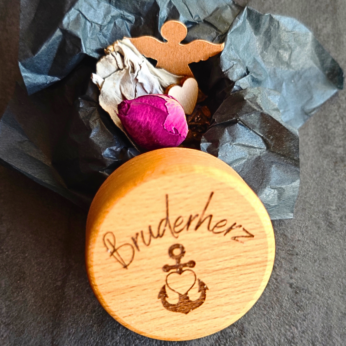 Geöffnete Mementovia-Box "Stille Umarmung" mit den symbolischen Beigaben Lavendel, Rose, Salbei, Engel und Herz – sanfte Begleiter in der Trauer. Im Vordergrund der Deckel der Box mit dem Motiv "Glaube, Liebe, Hoffnung" und dem gravierten Wort "Bruderherz" - Die Beigaben liegen auf schwarzem Seidenpapier.